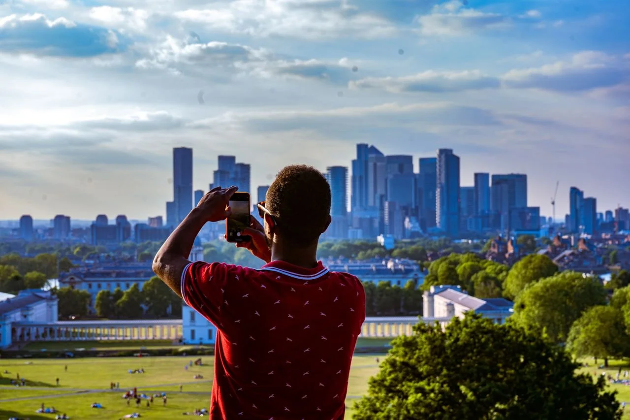 Person in red shirt taking a photo of a city skyline with tall buildings and skyscrapers, park with trees and people in the foreground.