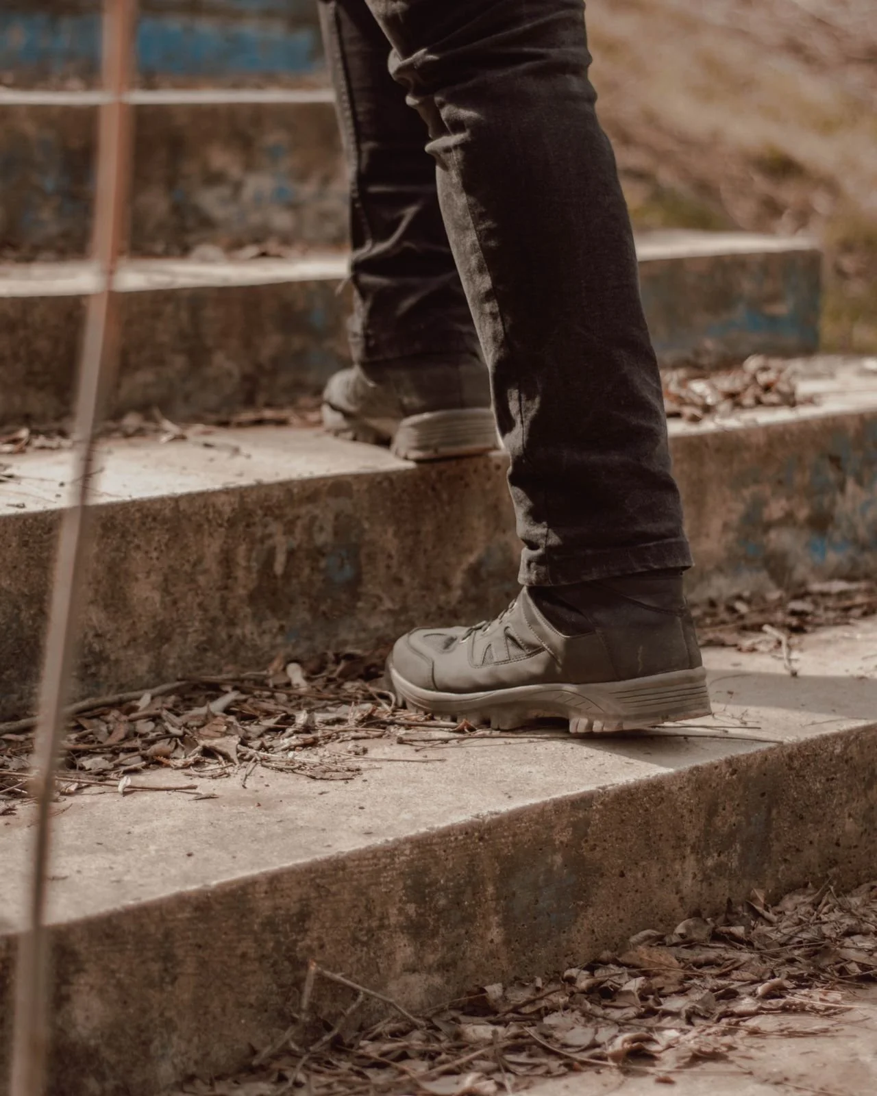 Close-up of a person wearing black jeans and gray sneakers walking up outdoor concrete stairs covered with dry leaves, with a metal railing on the left side.