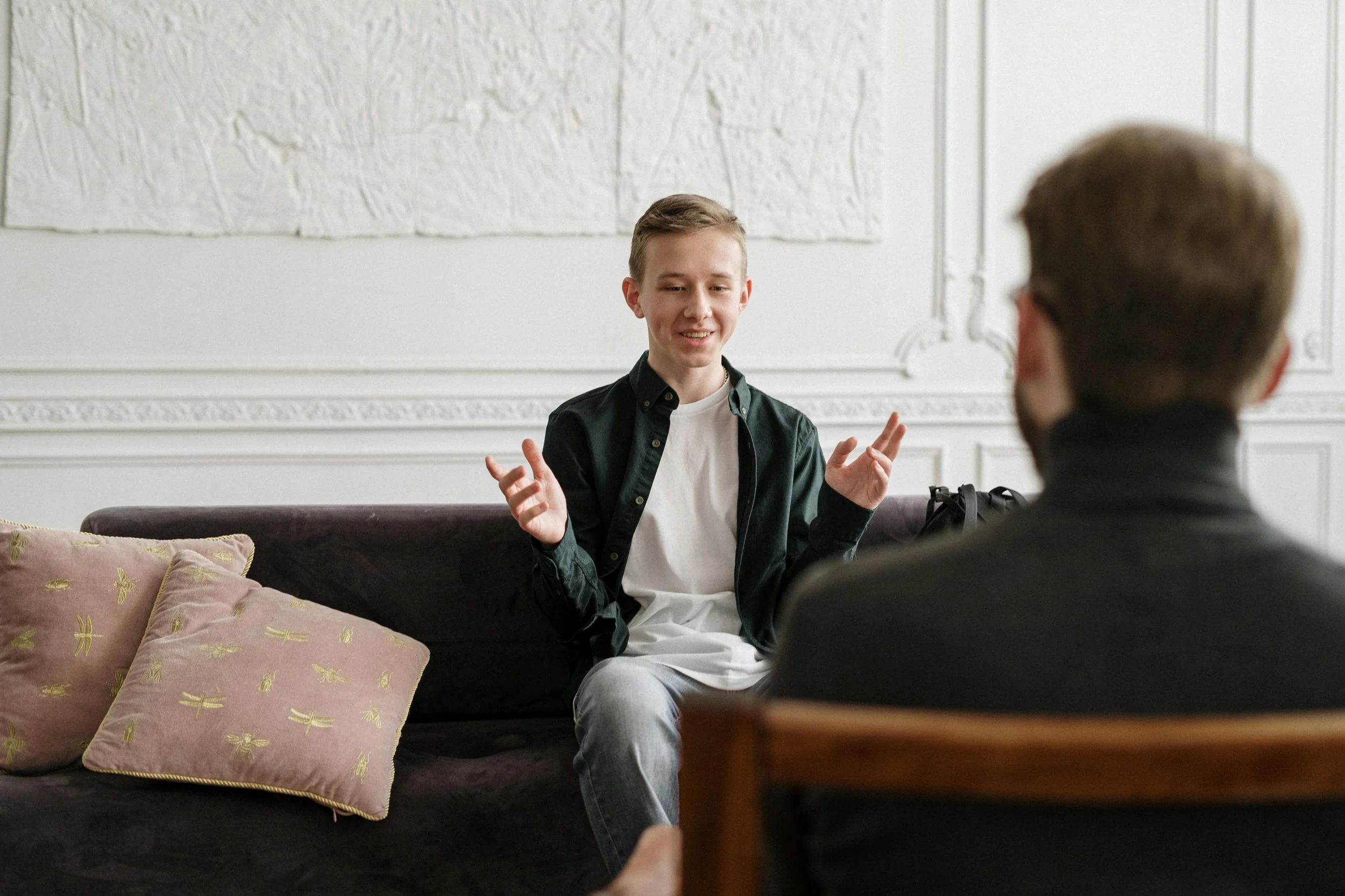 Two young men are sitting and talking in a living room with white walls. One man is facing the camera, smiling and gesturing with his hands, while the other man has his back to the camera. There are pink pillows with gold dragonfly patterns on a dark sofa.
