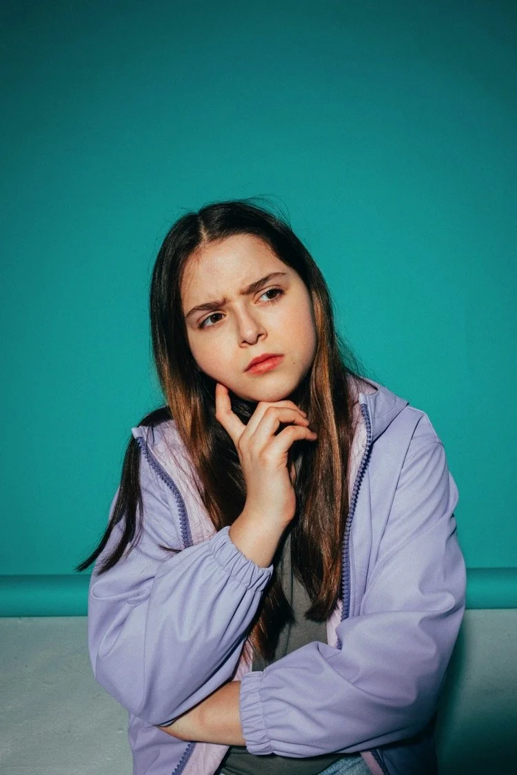 A young girl with long brown hair, wearing a light purple jacket, sitting with her hand on her chin and a thoughtful expression on her face against a teal background.