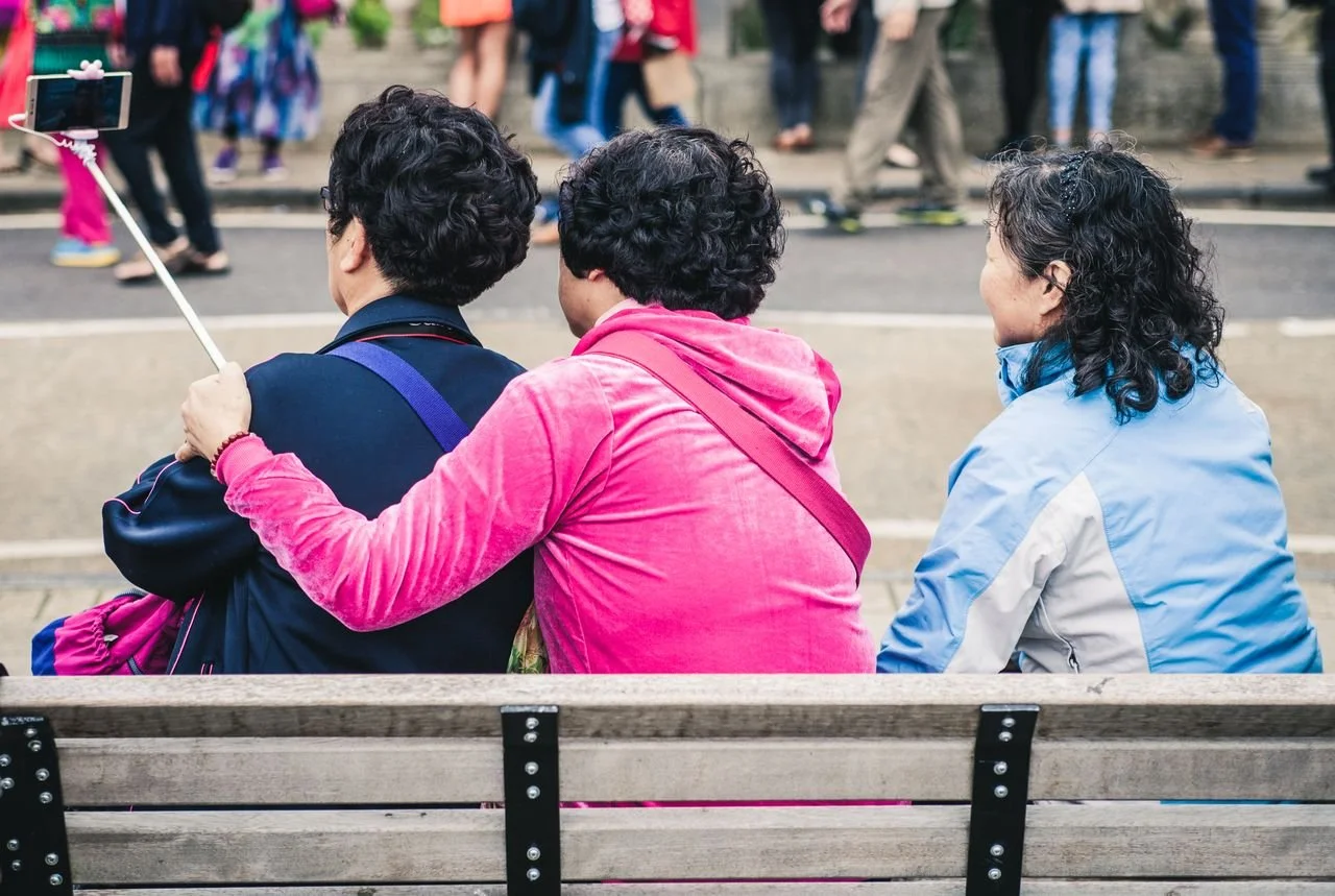 Three women sitting on a park bench, one with her arm around the other, taking a selfie with a smartphone on a selfie stick.