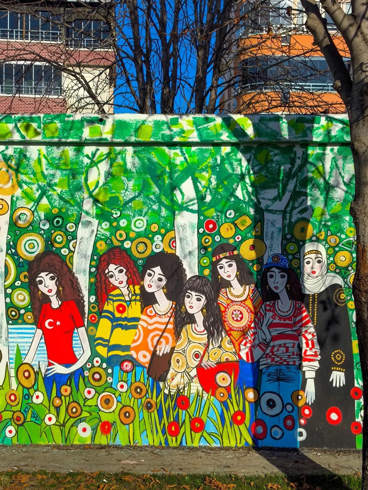 A colorful mural featuring six women with various hairstyles and traditional clothing patterns, standing in a field of flowers, with trees and modern buildings in the background.