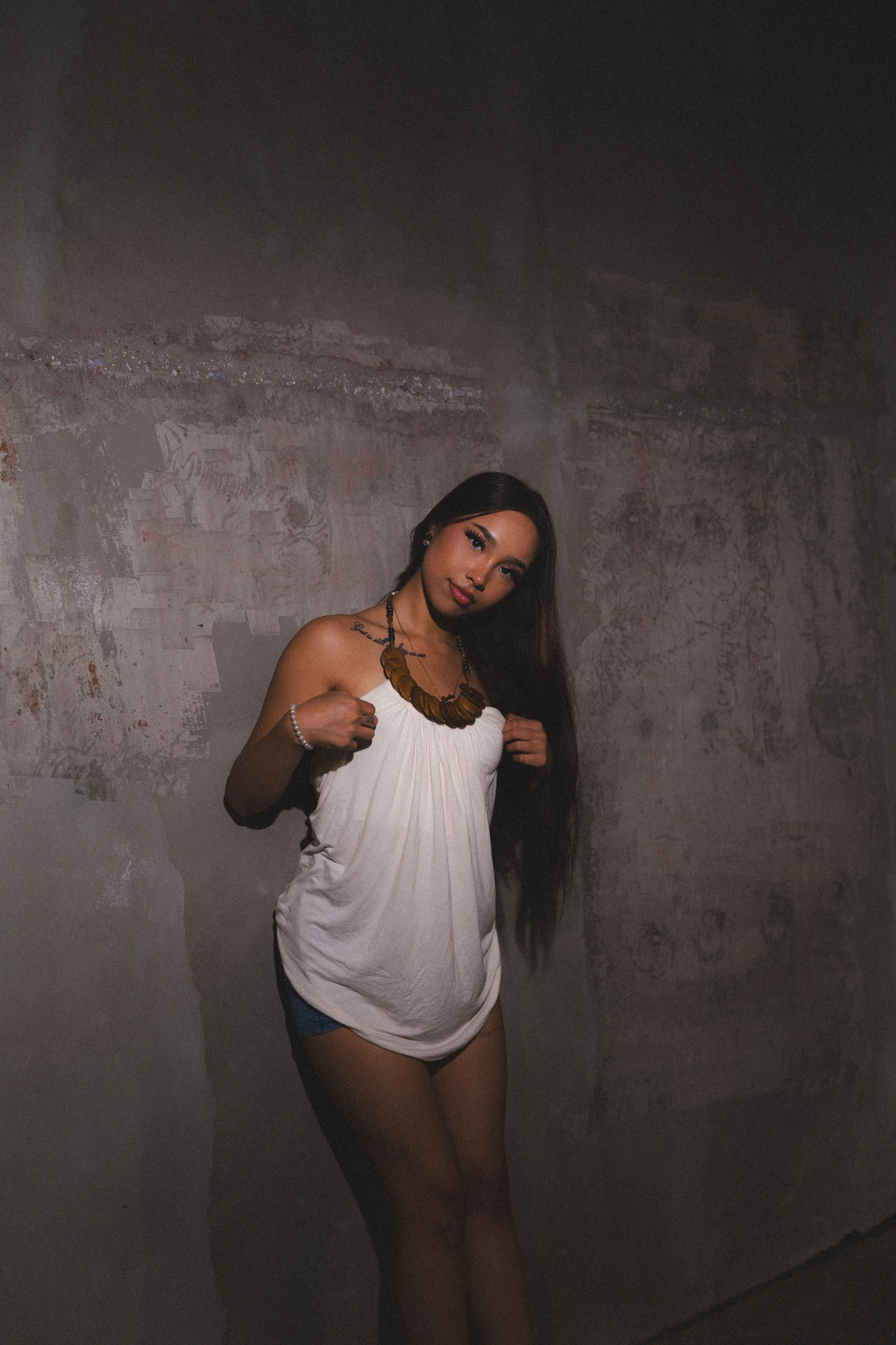 A woman with long dark hair wearing a white strapless top, dark shorts, a large wooden necklace, bracelet, and earrings, standing against a grungy gray wall.