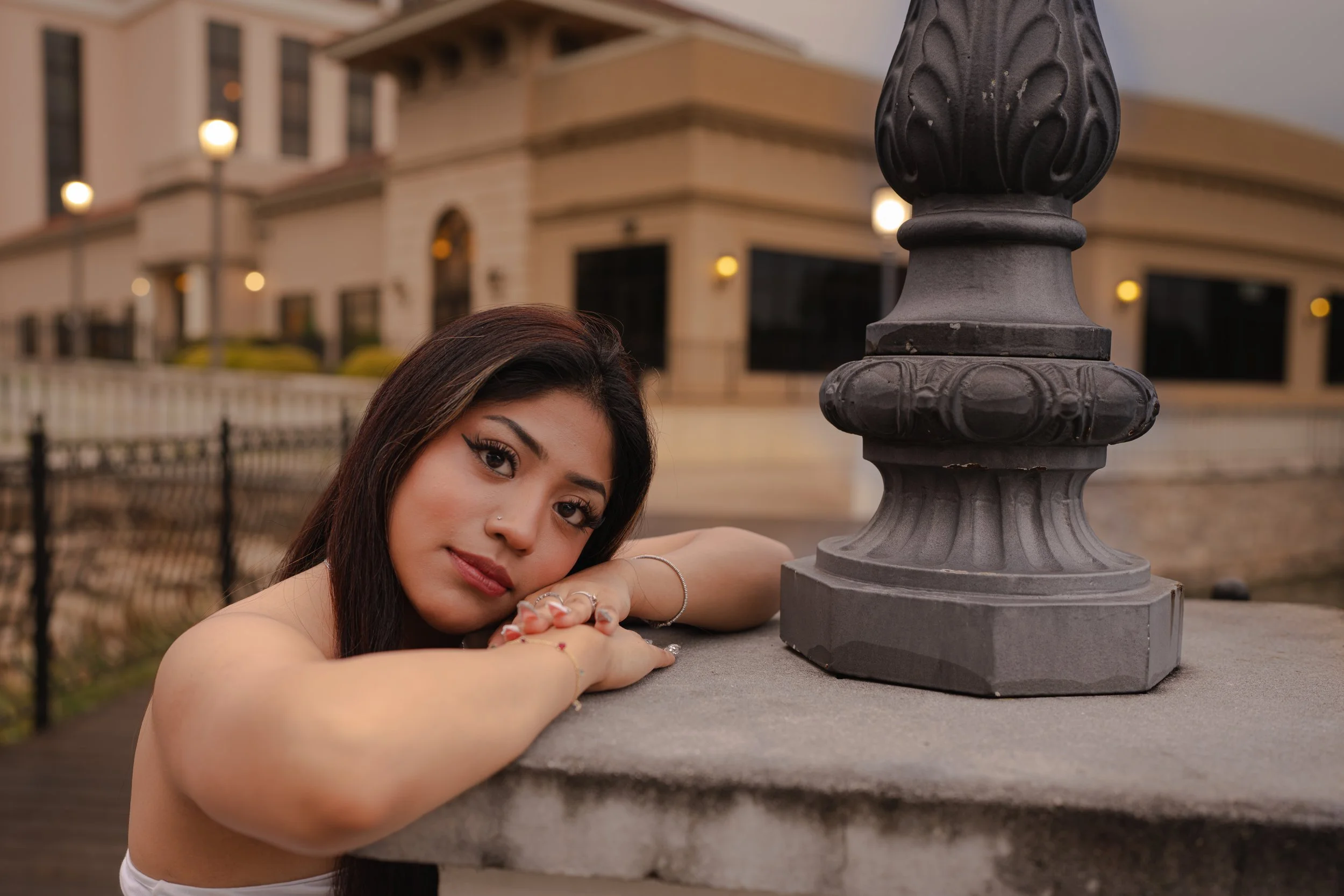 A woman with long dark hair and light skin leaning on a stone railing with her arms crossed, posing near a vintage-style black lamppost outside in front of a building at dusk.