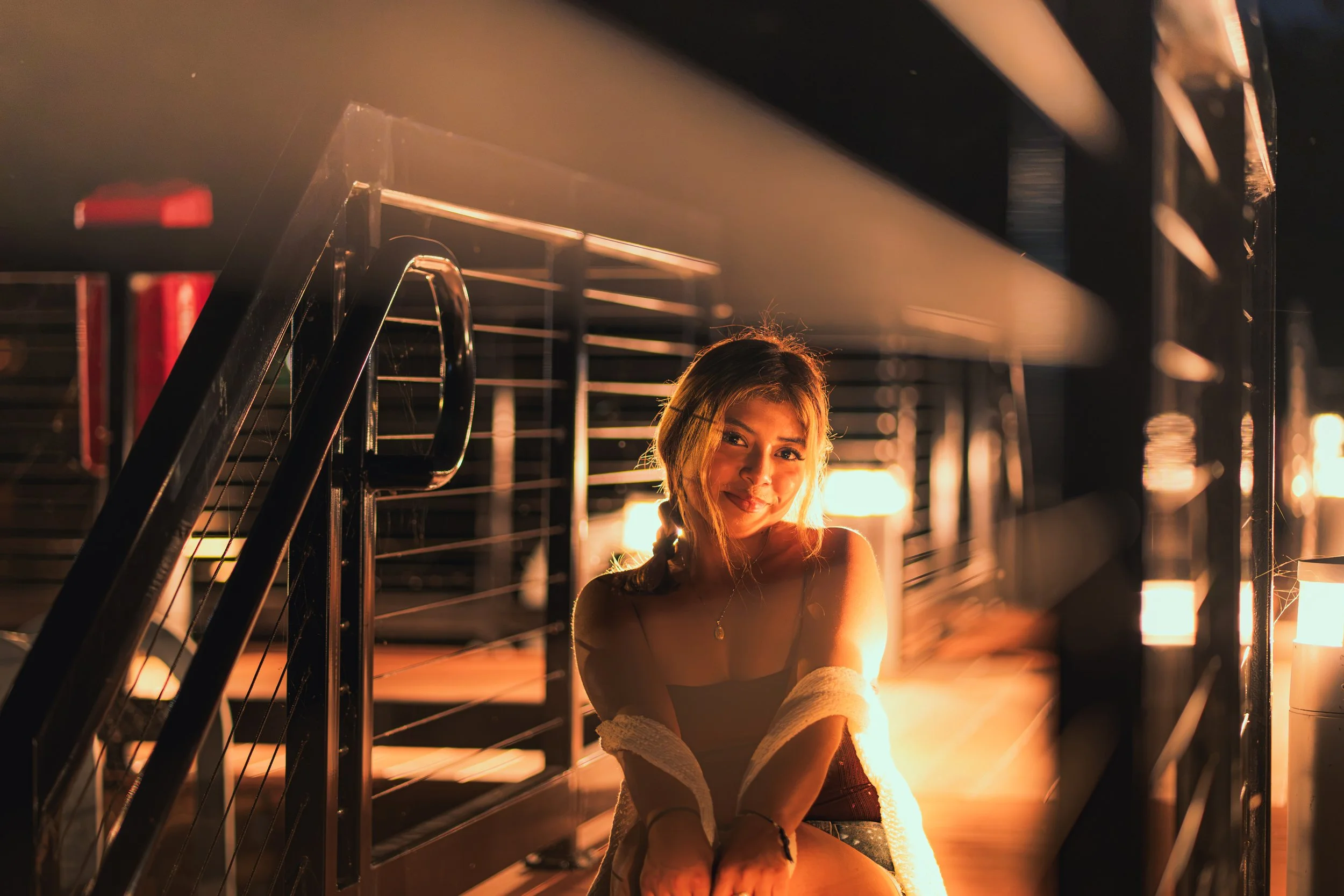 A young woman sitting on the floor next to a black metal staircase bathed in warm, orange backlight.