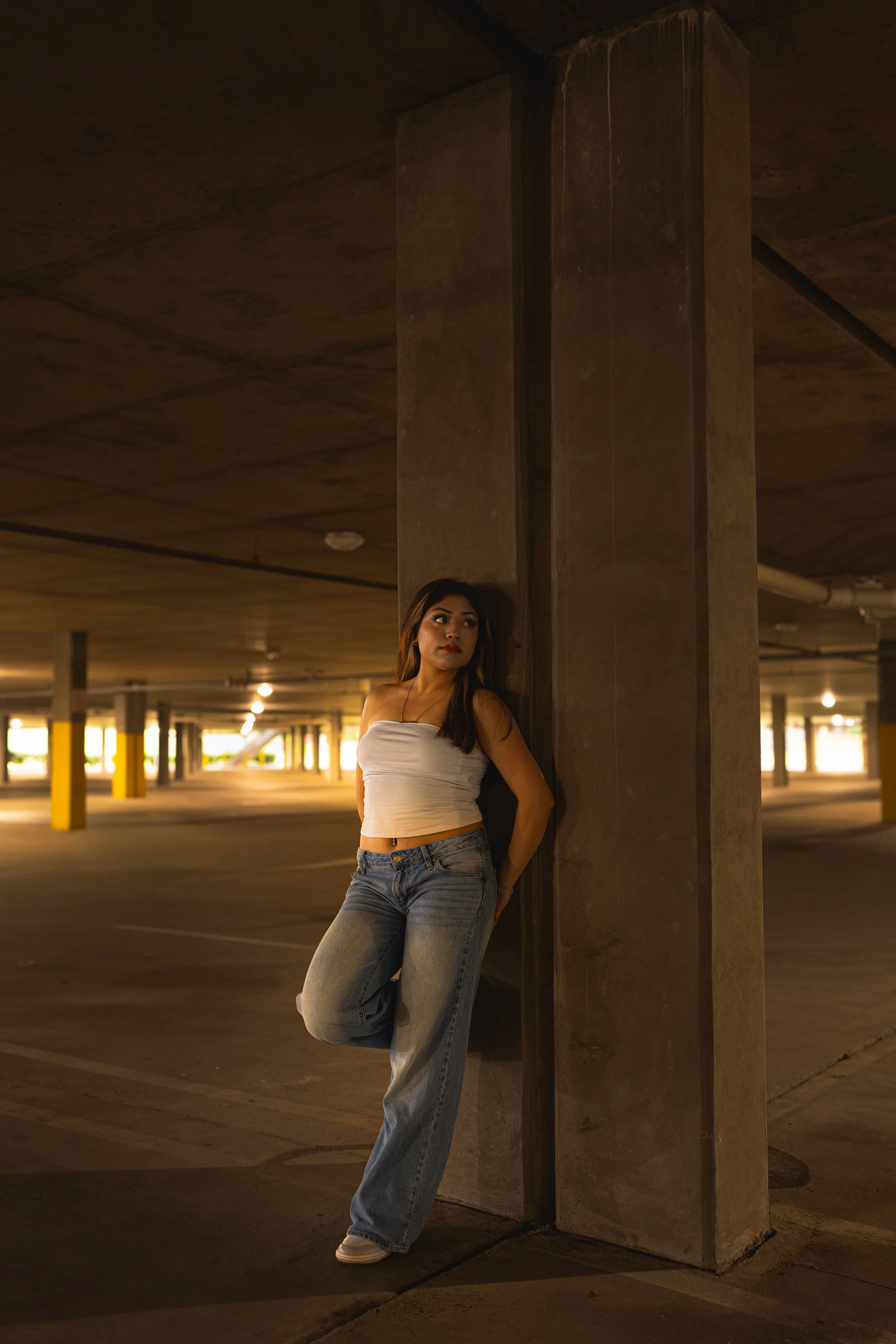A young woman in a white strapless top and blue jeans stands leaning against a concrete pillar in a parking garage. The background features lit parking spaces and concrete ceiling, with a warm, ambient lighting.