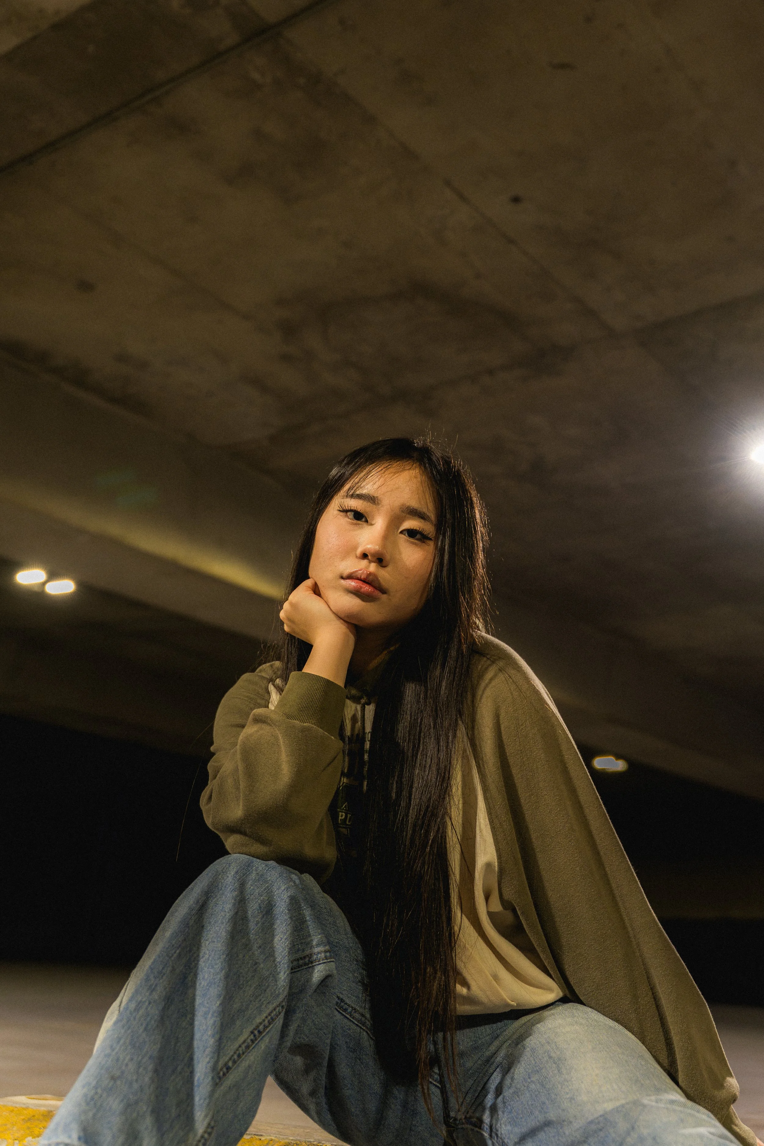 A young woman with long dark hair, wearing a khaki hoodie and jeans, sitting with one leg bent under her, in an underground parking garage with a concrete ceiling and overhead lighting.