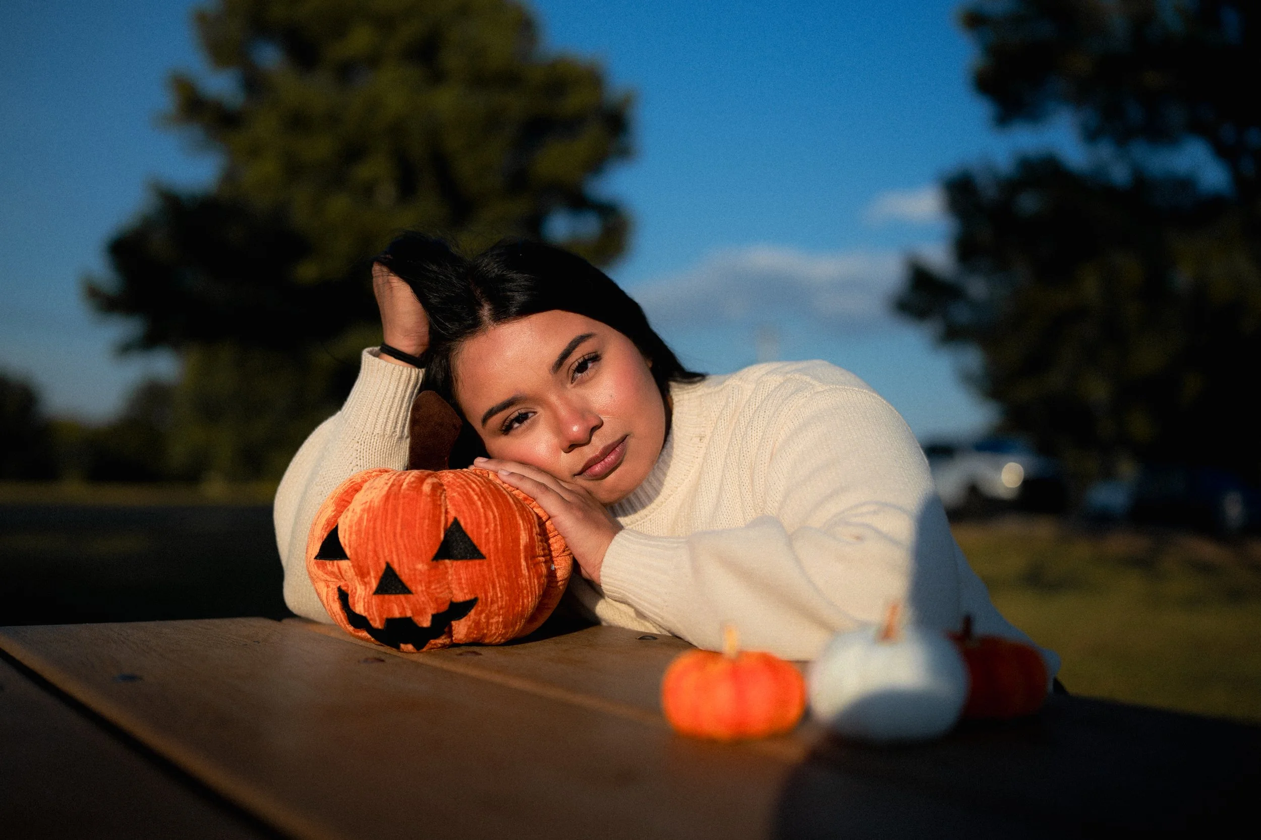A young woman lying on a table at sunset, resting her head on her folded arms next to a carved pumpkin with a jack-o'-lantern face, surrounded by small orange pumpkin decorations.