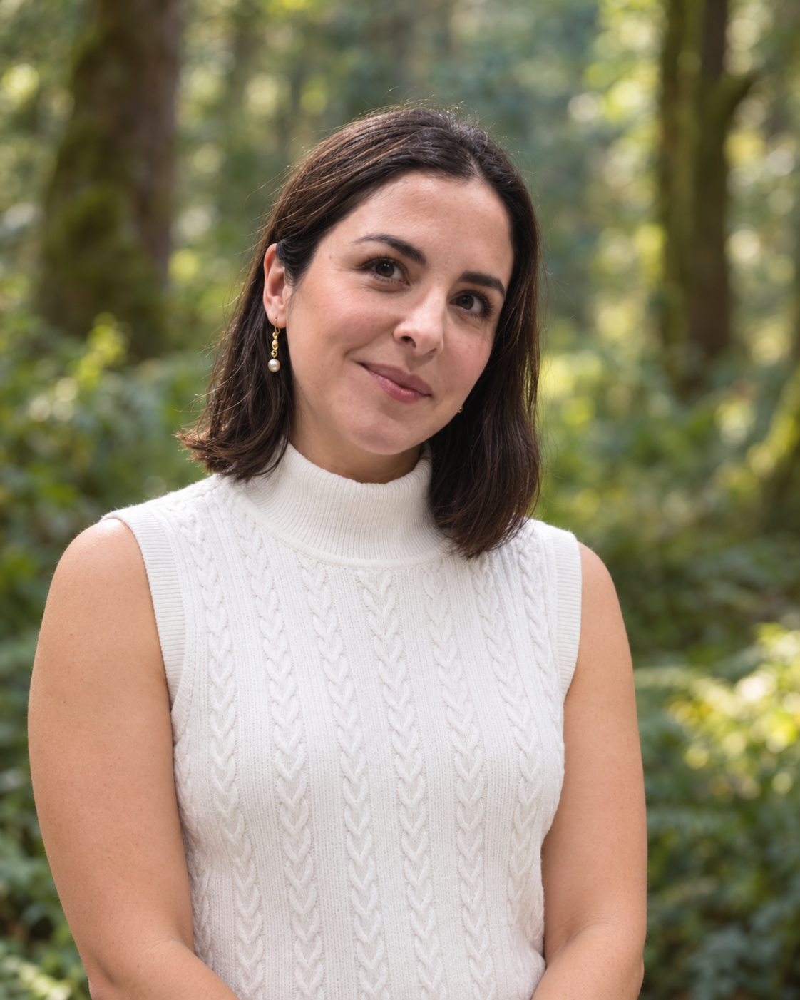 A woman with shoulder-length dark hair wearing a white sleeveless knitted turtleneck top and pearl earrings, standing outdoors in a lush green forest.