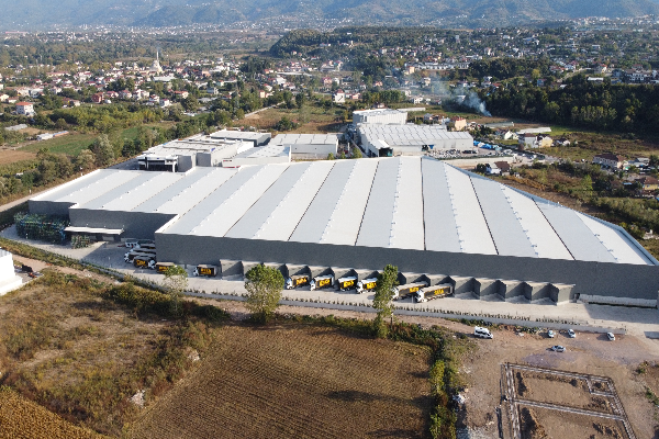 Large industrial warehouse with a curved roof, surrounded by trucks, trees, and open land, with a cityscape and mountains in the background.