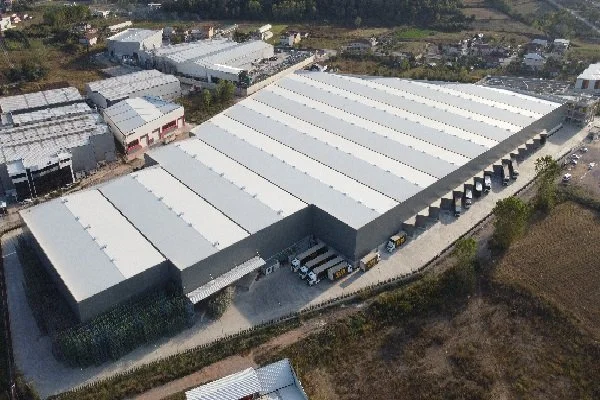Aerial view of a large industrial warehouse with white roof panels, surrounded by a paved lot and some parked trucks.