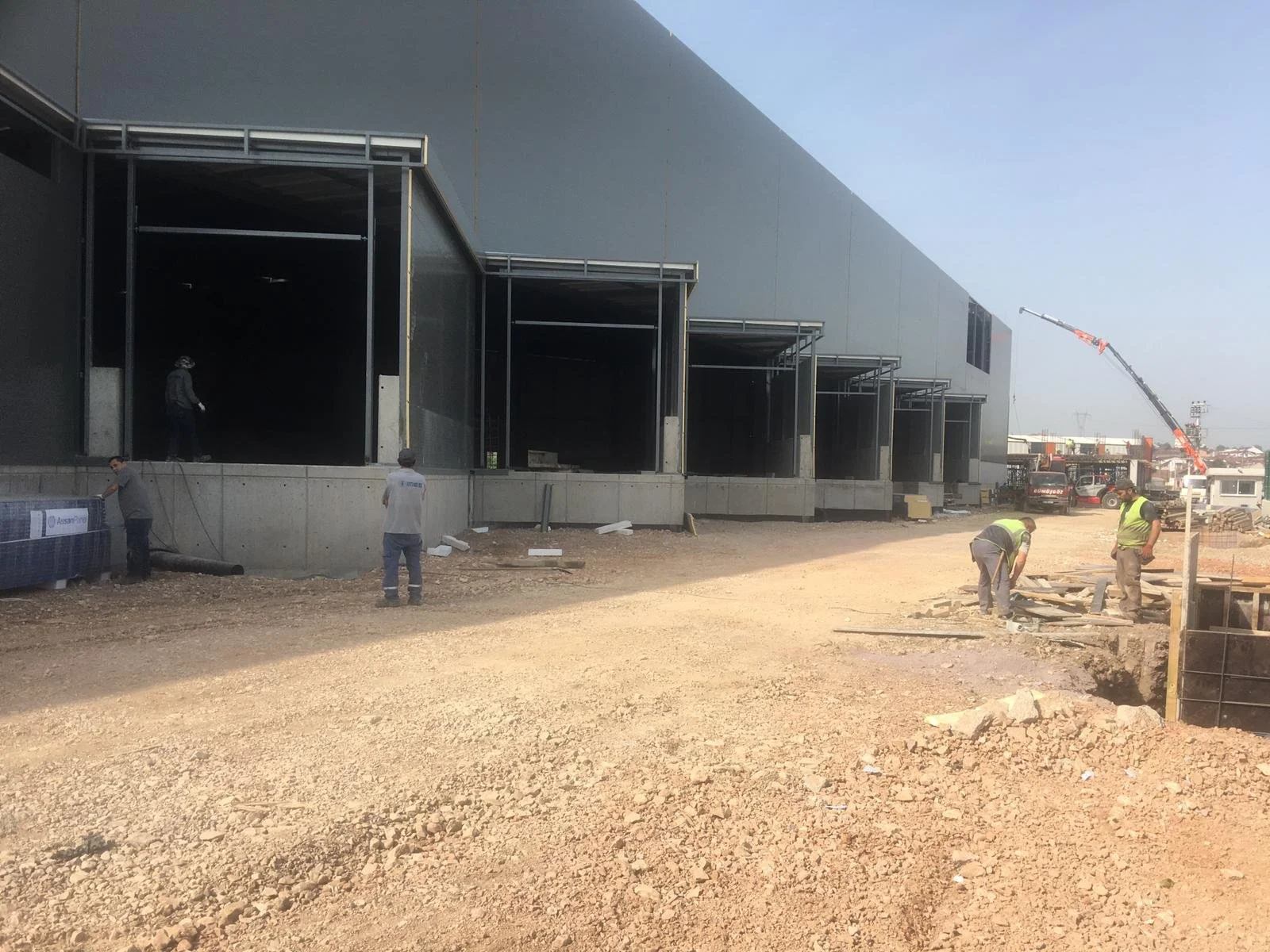Construction site with workers installing or constructing a large building, with a crane in the background and workers working on the ground.