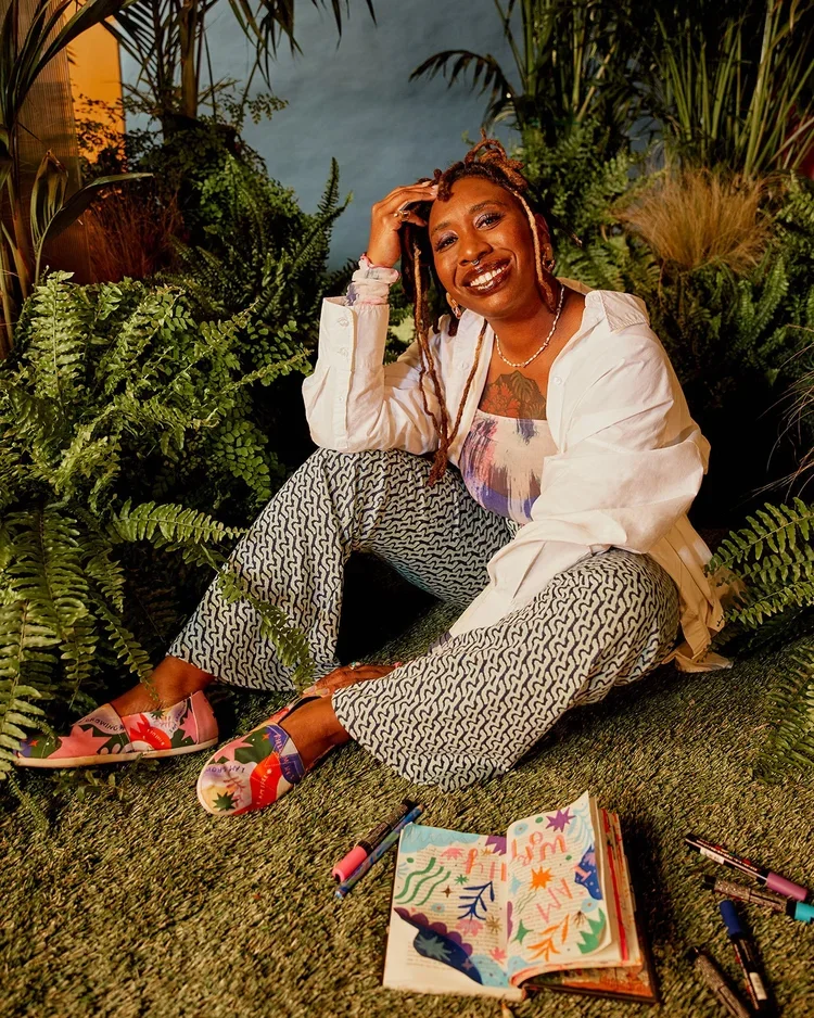 A woman sitting on green carpet surrounded by lush green plants, wearing patterned pants, colorful shoes, a white shirt, and a colorful top, smiling and posing with her hand in her hair. There are markers and a colorful open notebook nearby.