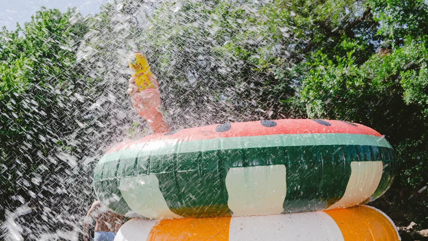 A person holding a yellow Slurpee cup on top of an inflatable water tube, surrounded by water spray outdoors with green foliage in the background.