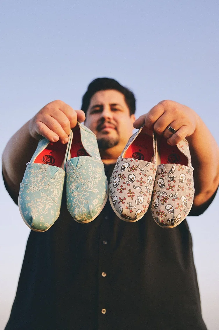 A man holding up two pairs of colorful slippers with festive patterns, one light blue with white designs, and the other white with skulls, bones, and holly, against a clear sky background.