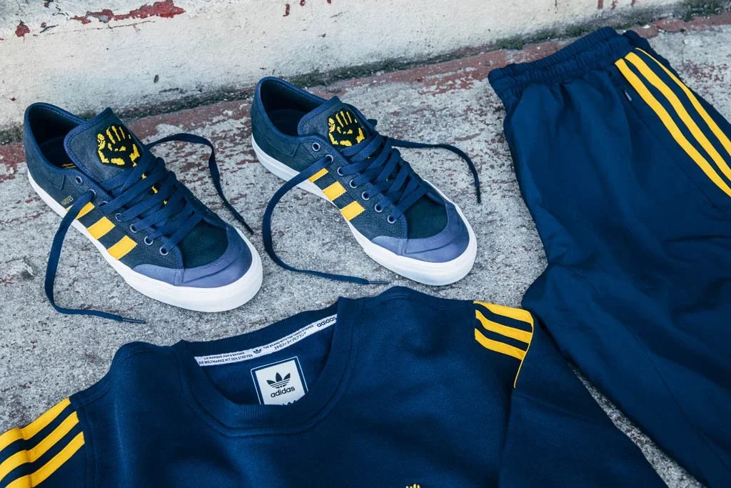 A pair of blue Adidas sneakers with yellow three stripes and white soles, a blue Adidas sweatshirt with yellow stripes on the sleeves, and matching blue Adidas pants with yellow stripes, all placed on a concrete surface against a white wall.