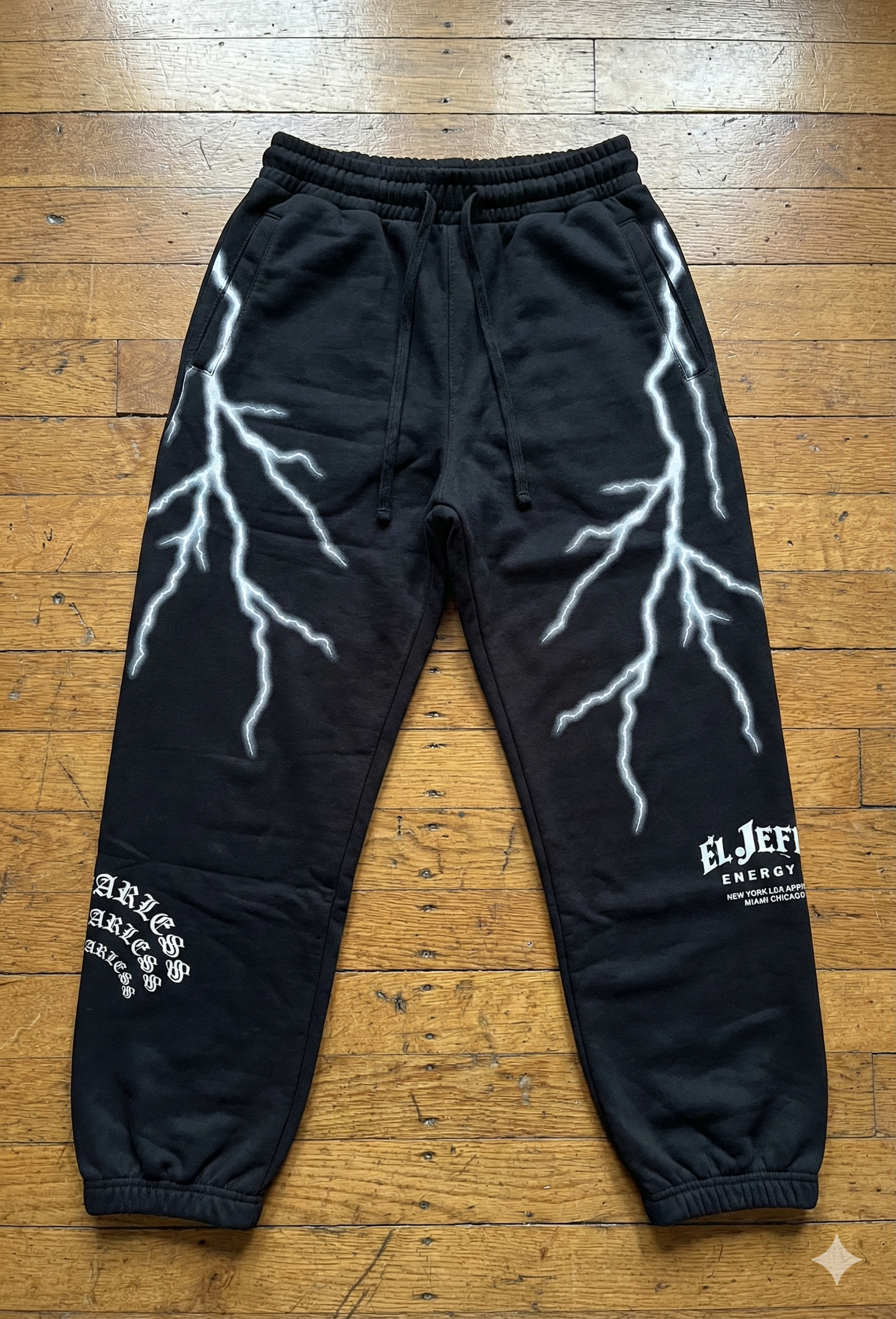 Black sweatpants with lightning bolt designs on the thighs and logos, laid on a wooden floor.