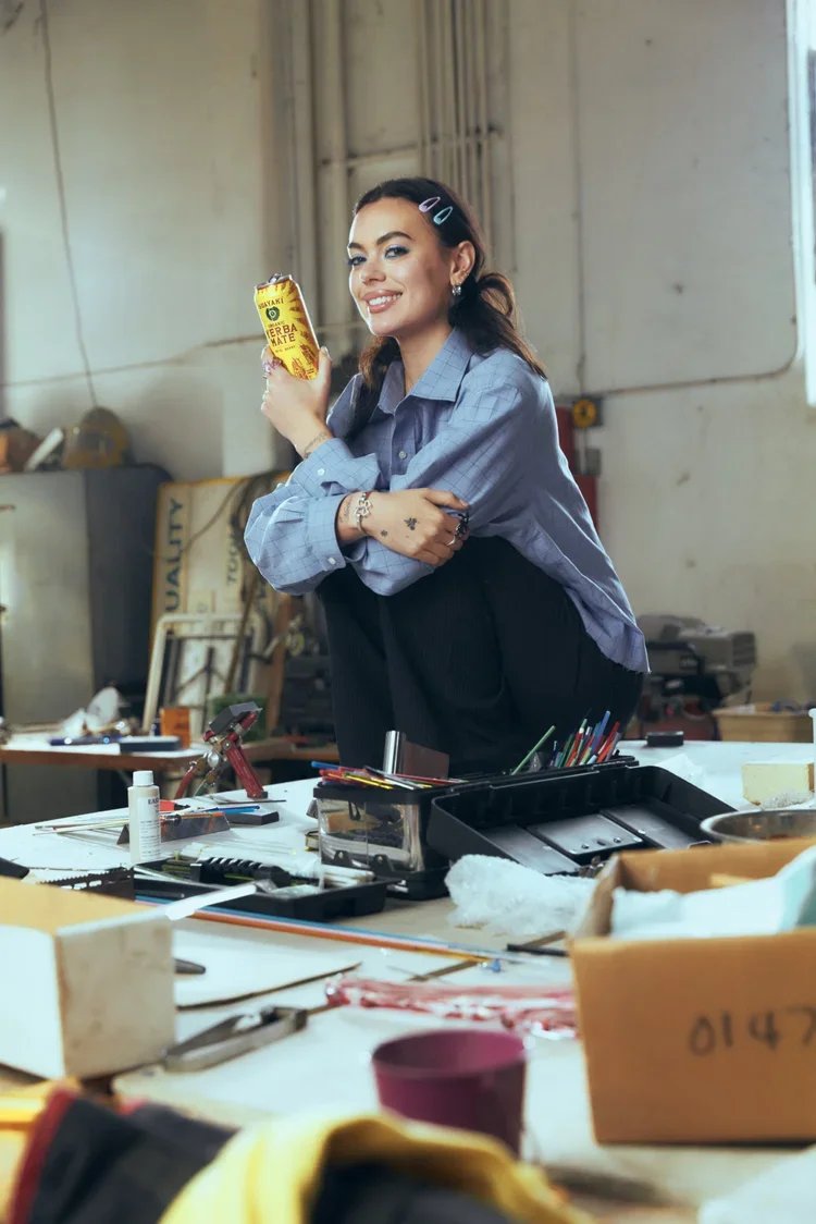 A woman with dark hair, wearing a blue button-up shirt and black pants, is smiling and holding a yellow can of Kurkure herbal snack in an industrial workspace with cluttered tables and tools.