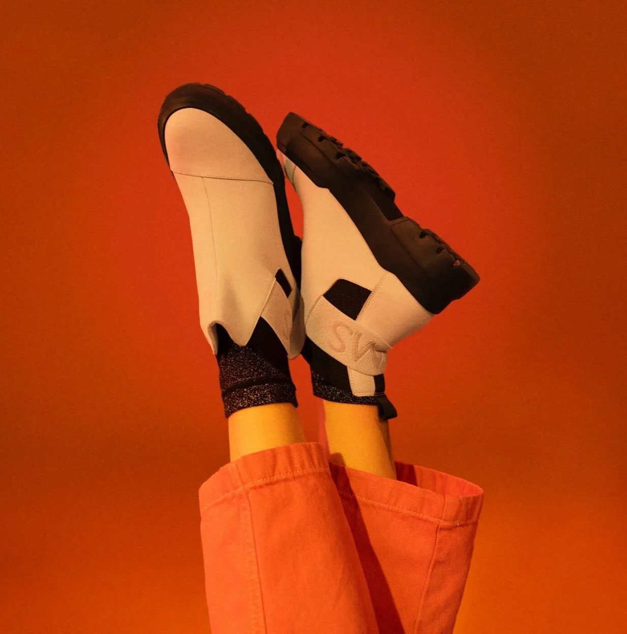 A person wearing white and black sneakers and black socks, with orange pants, against an orange background, with legs raised in the air.