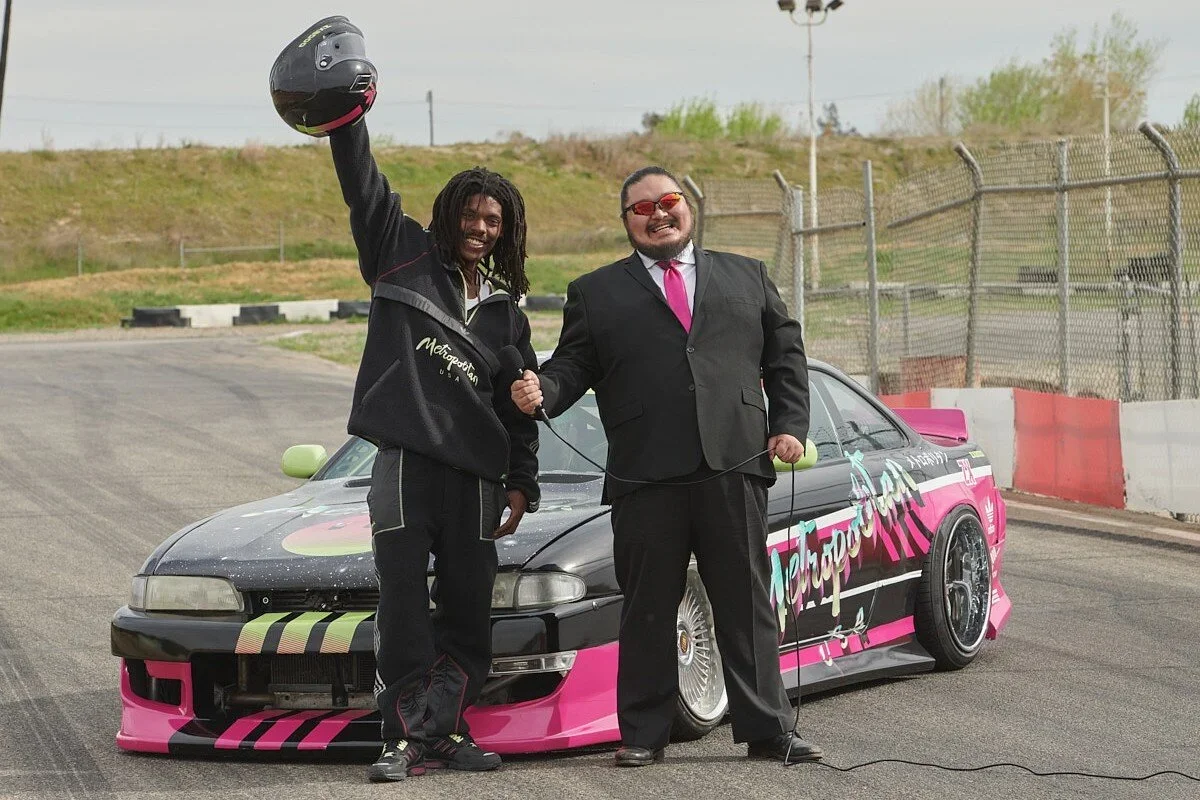 Two men standing in front of a race car on a track, one man wearing a racing suit and holding a helmet, the other in a black suit with a pink tie, both smiling and holding hands, during daytime.