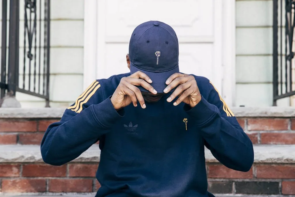 Person wearing a navy blue cap and tracksuit with gold stripes on the sleeves, sitting on brick steps, with head bowed and hands adjusting the cap, in front of a house with a white door and black railings.
