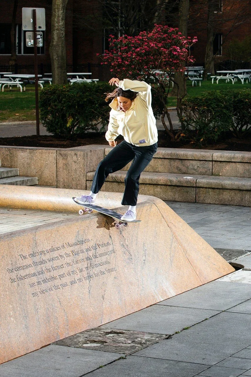 A woman skateboarding on a ledge at a park during evening with trees, bushes, and a blooming pink shrub in the background.