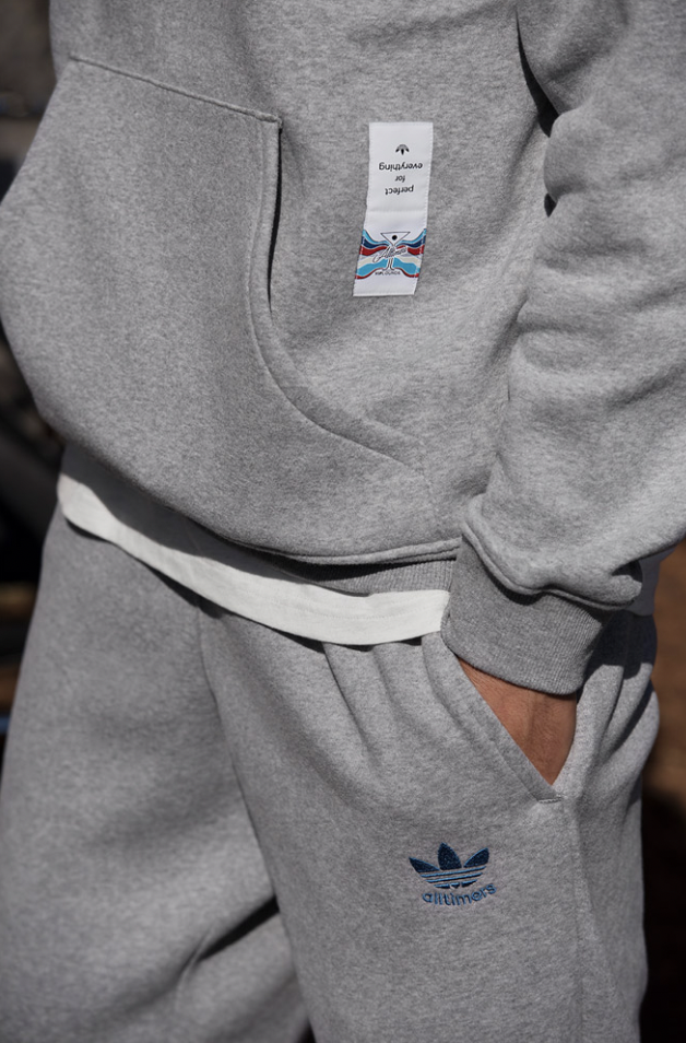 Close-up of a person wearing a gray hoodie and matching sweatpants, with a hand in the pocket of the sweatpants. The hoodie has a white tag hanging from the pocket, and the sweatpants have a blue Adidas logo with 'athleisure' written underneath.