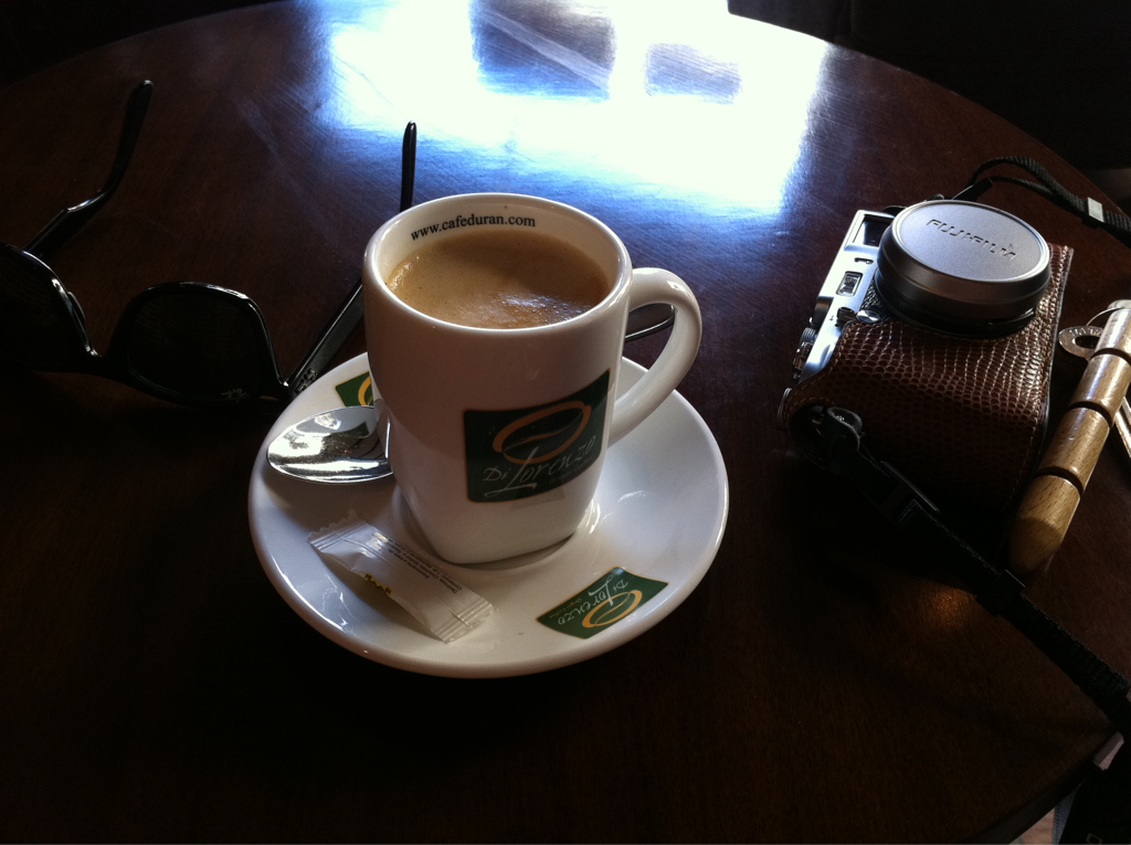 A white coffee cup filled with coffee, placed on a saucer with a spoon and sugar packet, two sunglasses, a camera with a lens cap, and a wooden wind instrument on a dark wooden table.