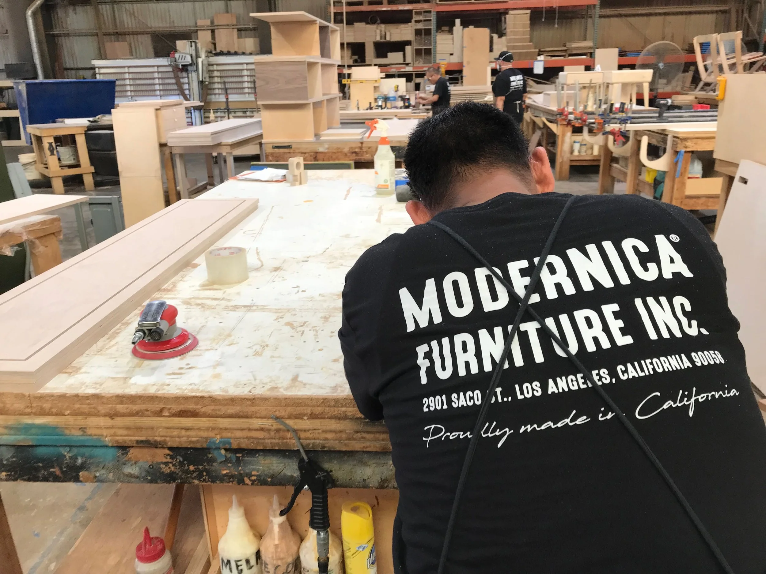 Inside a woodworking shop with shelves of wood, tools, and furniture projects. A person wearing a black shirt with white text on the back is working at a large workbench. The person’s shirt features the words "MODERNICA FURNITURE INC." along with an 