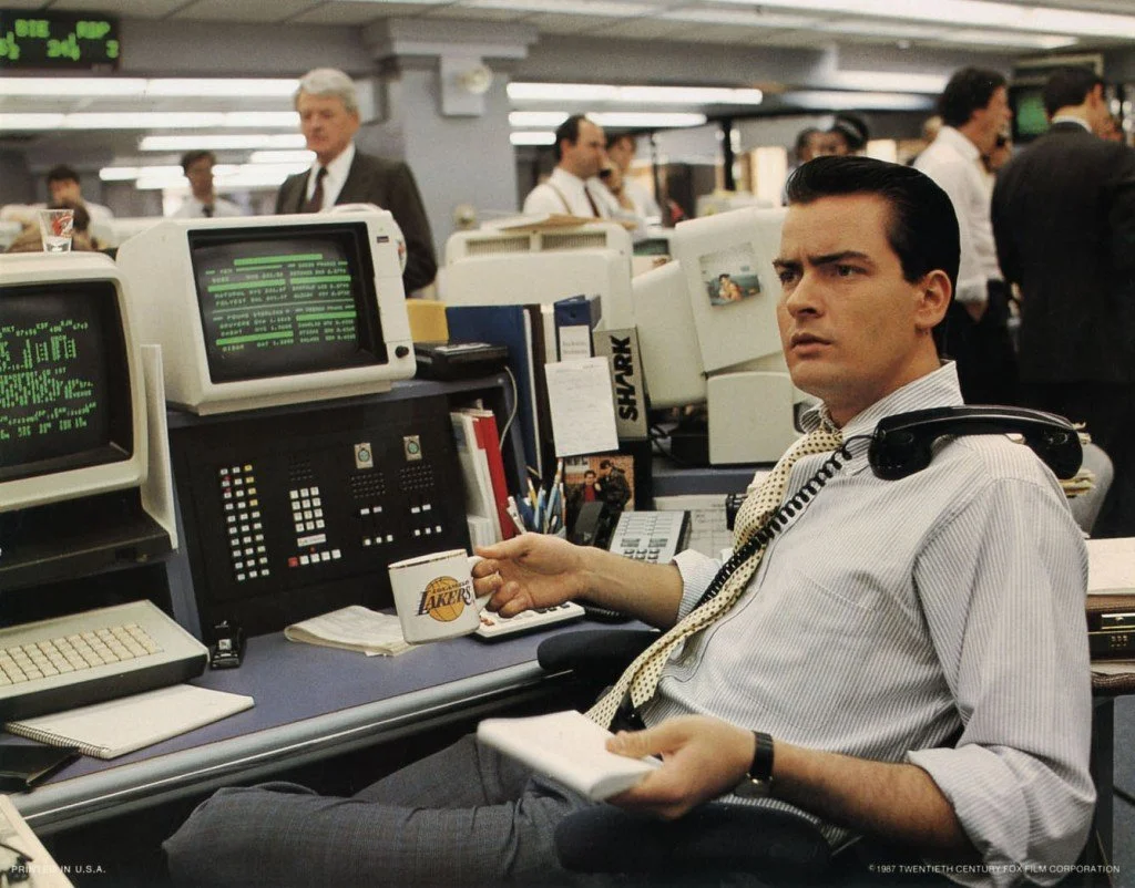 A man with dark hair, wearing a white dress shirt, is sitting at a cluttered office desk with multiple computers and monitors, holding a Lakers mug, with office colleagues working in the background.