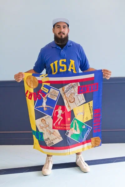 A man with a beard wearing a blue USA sweatshirt and a gray cap holding a colorful quilt with Olympic-themed patches.