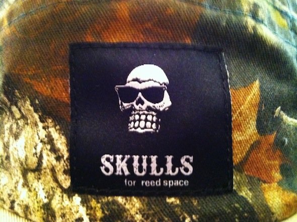 Clothing label with a skull wearing sunglasses, with the text 'Skulls for Reed Space' on a camouflage background.