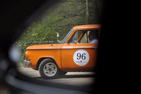 Orange vintage car with a round white sticker numbered 96 on the side, parked outdoors near trees.