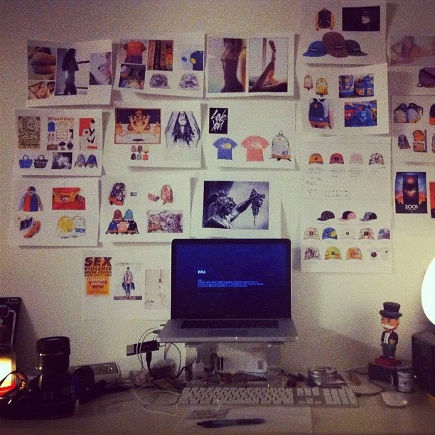 A desk with a laptop, keyboard, and various objects. The wall behind the desk has numerous photos, drawings, and posters pinned up, including images of caps, clothes, and abstract art.