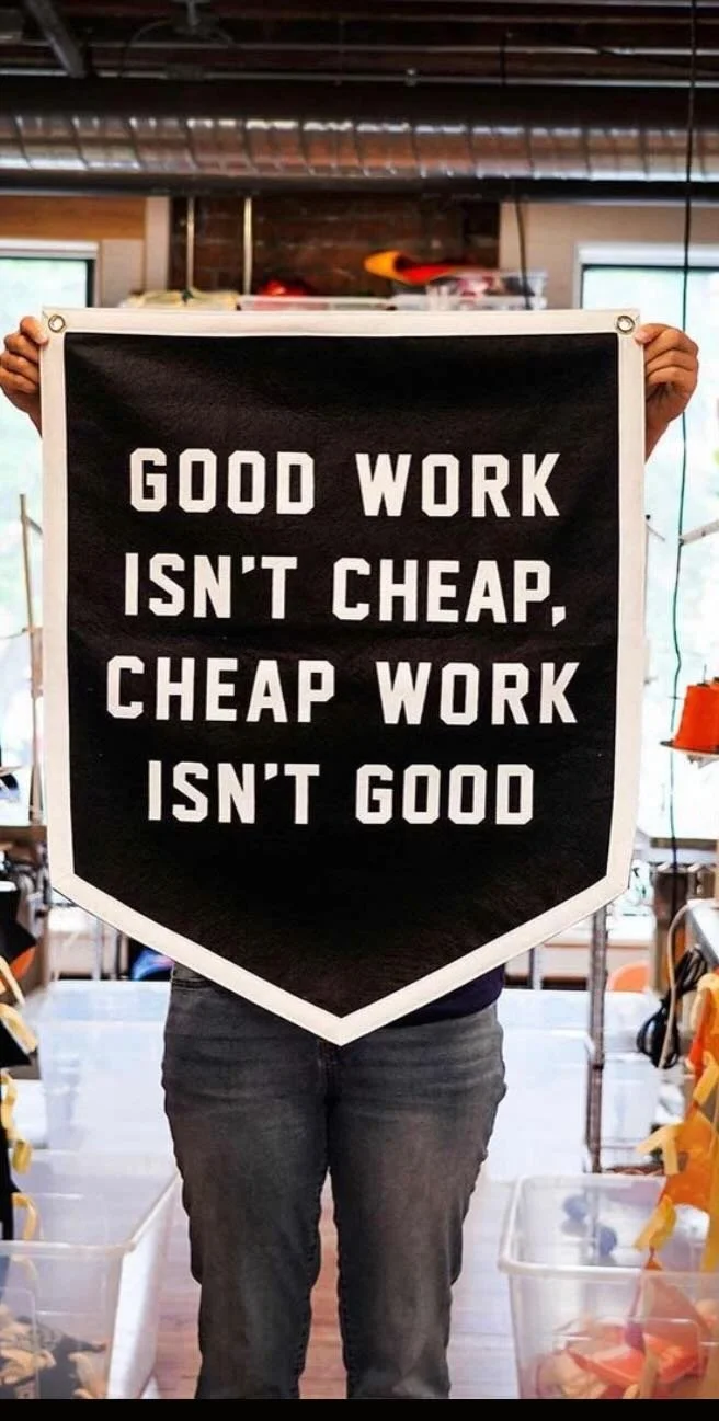 Person holding a black and white sign that reads, "Good work isn't cheap, cheap work isn't good," inside a room with industrial ceiling and windows.