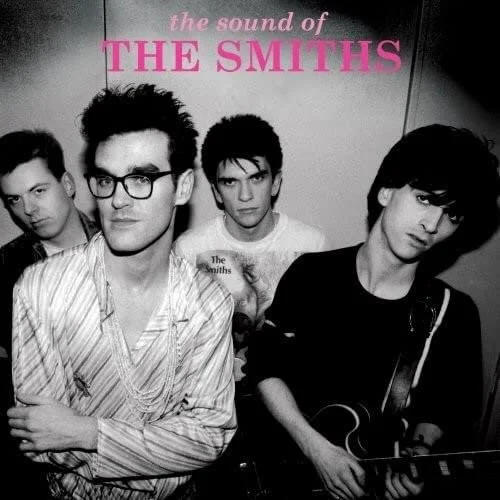 Black and white photo of the band The Smiths with four members, one holding a guitar, in front of a wall with a sign that reads 'the sound of THE SMITHS' in pink text.