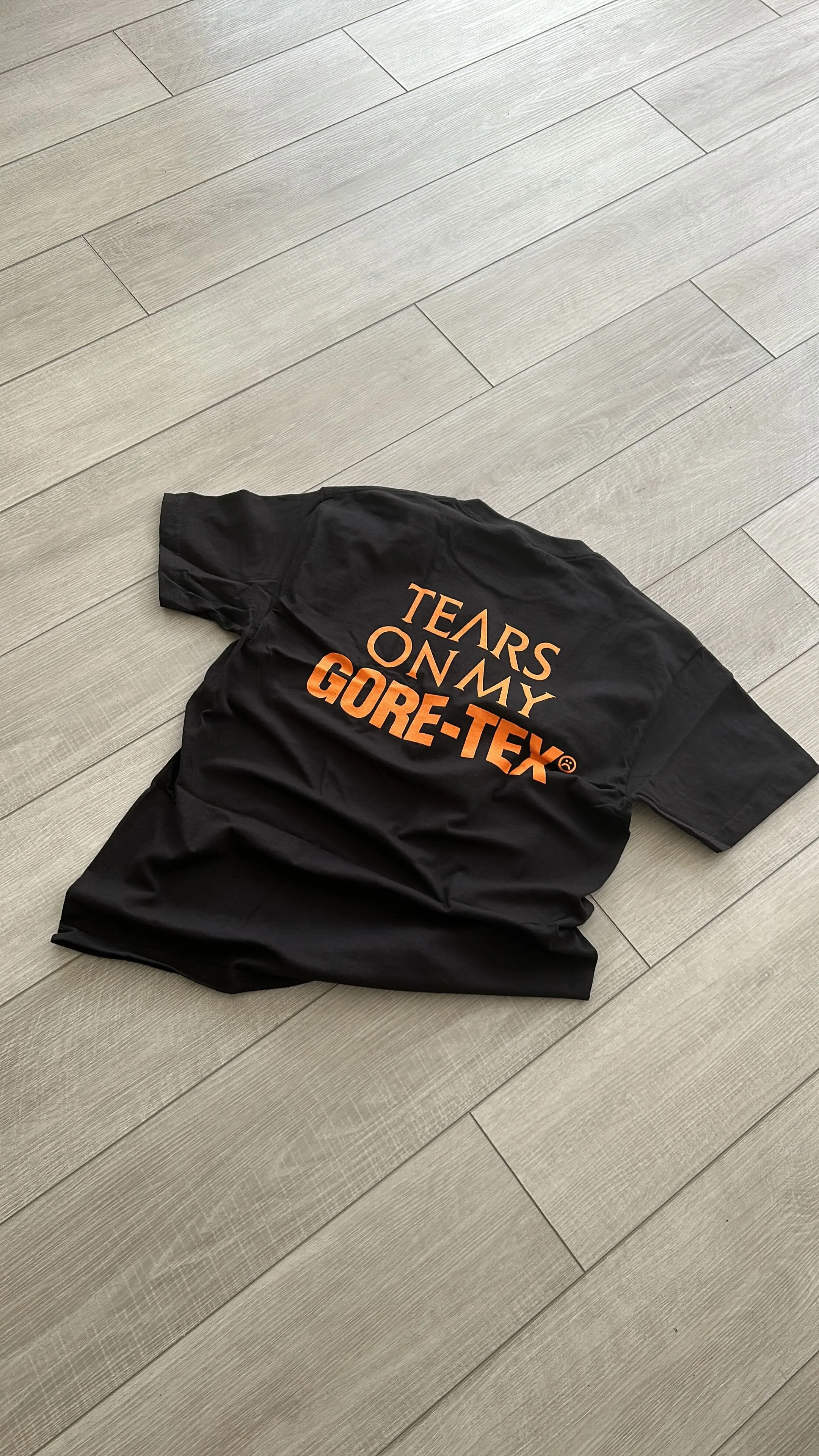 Black GORE-TEX jacket with orange text reading 'TEARS ON MY GORE-TEX' lying on a light-colored wooden floor.