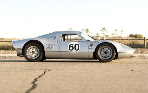 A vintage silver race car with the number 60 on its door, parked on a road with a desert landscape and palm trees in the background.