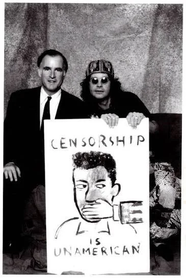 Two men sitting together, one in a suit and the other with sunglasses, holding a protest sign depicting a person with their mouth covered and a hand over their mouth, with the words 'CENSORSHIP UNAMERICAN'.