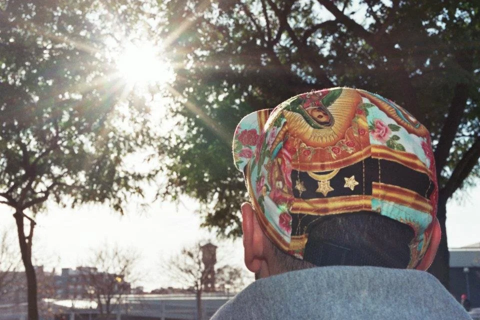 Back of a person wearing a colorful hat with Chinese embroidery design, sitting outdoors on a sunny day under a tree.