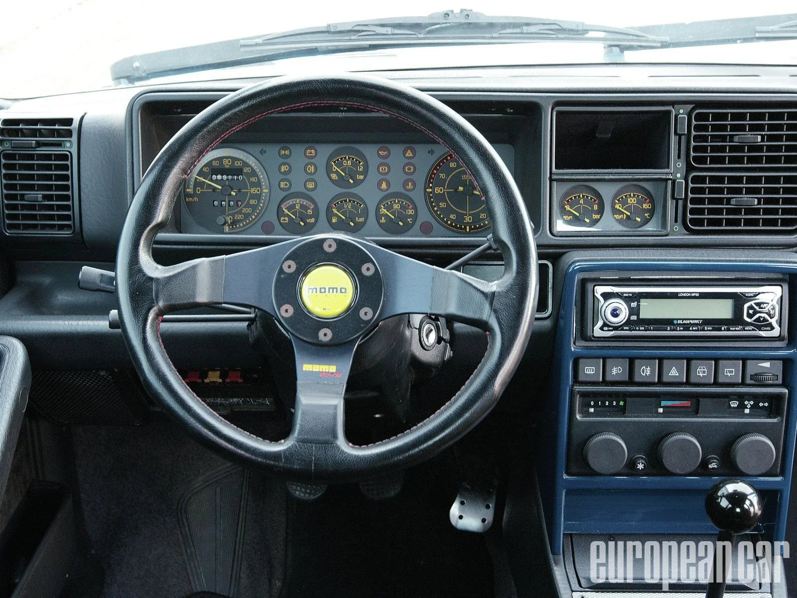 The interior of a vehicle showing a Momo racing steering wheel, a dashboard with multiple gauges, including speedometer, tachometer, and fuel gauge, and a Blaupunkt stereo system.