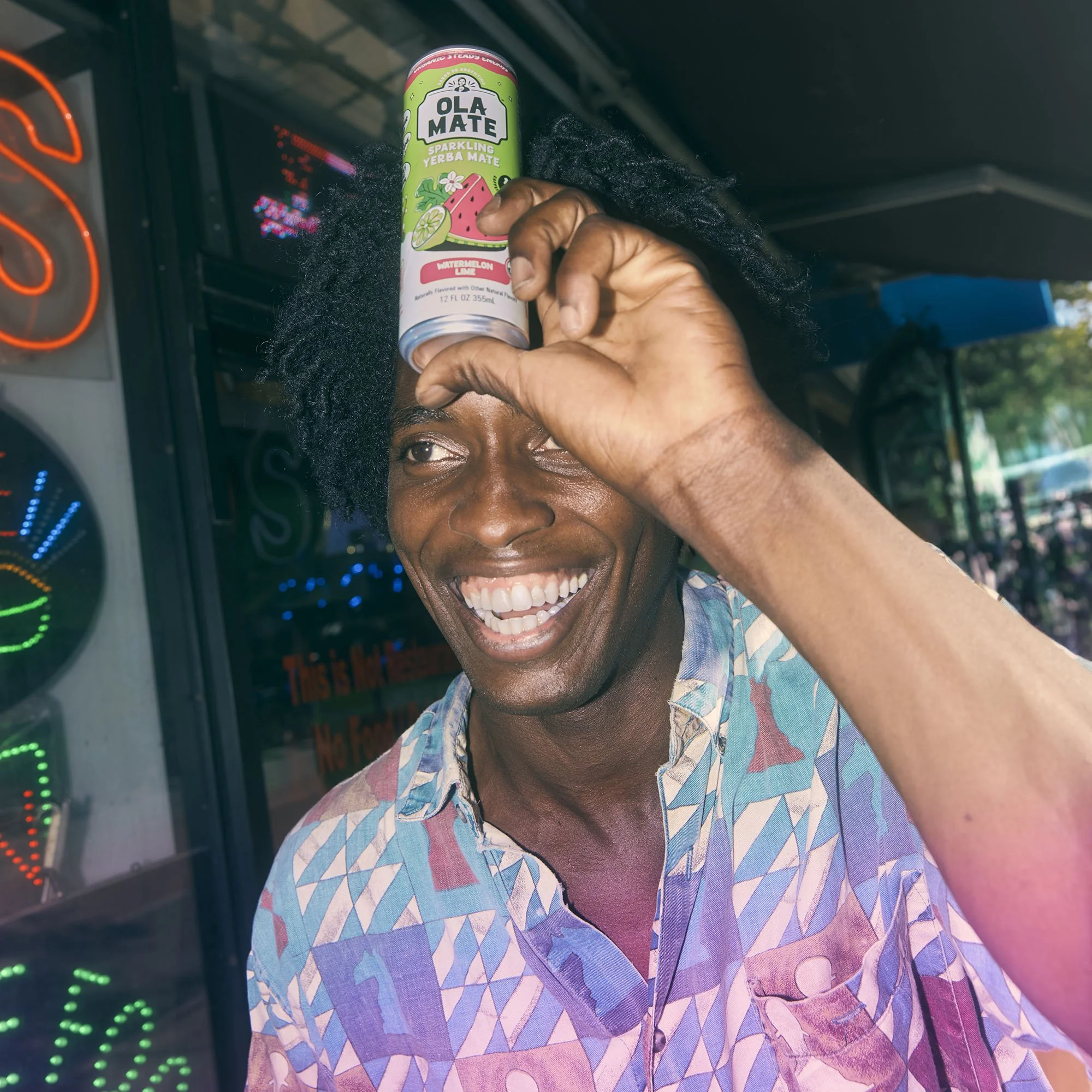 Person holding a can of Ola Mate Sparkling Yerba Mate watermelon lime flavor above their eye, smiling and wearing a colorful patterned shirt with black hair styled in dreadlocks.