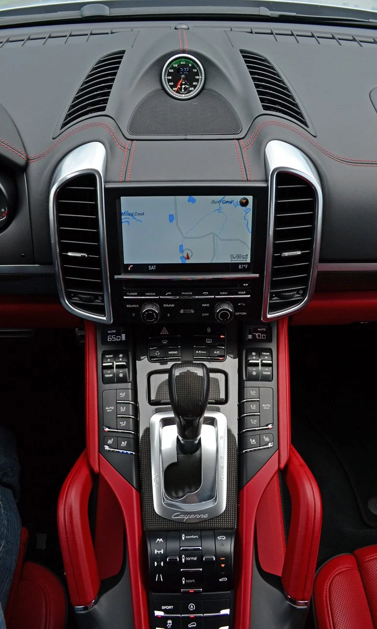 Interior view of a high-end sports car dashboard with a central touchscreen navigation system, gear shift, and various control buttons, featuring red accents and leather trim.