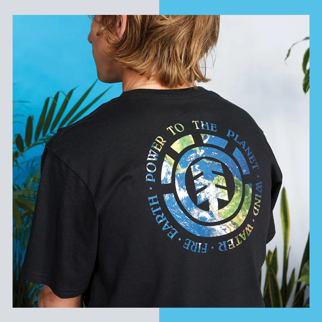 A person with blond hair is wearing a black T-shirt with a circular Earth-themed design on the back, featuring the Earth and the Earth Defense Force logo, along with the words 'Power to the Planet, Water, Earth, Fight, Free, Preserve' around the circ