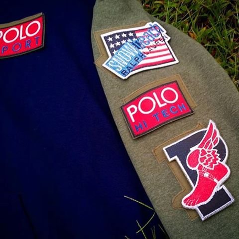 Close-up of a green fabric surface with three patches: American flag, POLO M TECH, and a mascot patch, next to a navy blue fabric with part of another POLO patch visible.
