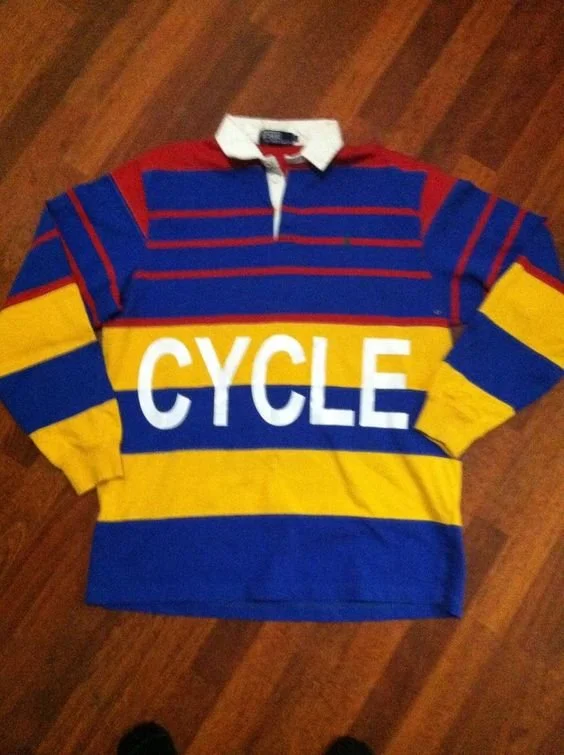 Colorful rugby shirt with red, blue, and yellow horizontal stripes and the word "CYCLE" printed on the front, laying flat on a wooden surface.