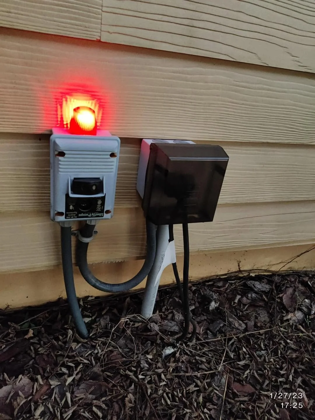 Outdoor view of electrical wiring and devices mounted on the side of a house with wooden siding. Includes a red light indicator, a white electrical box, and a black device, with wires running to ground among fallen leaves.