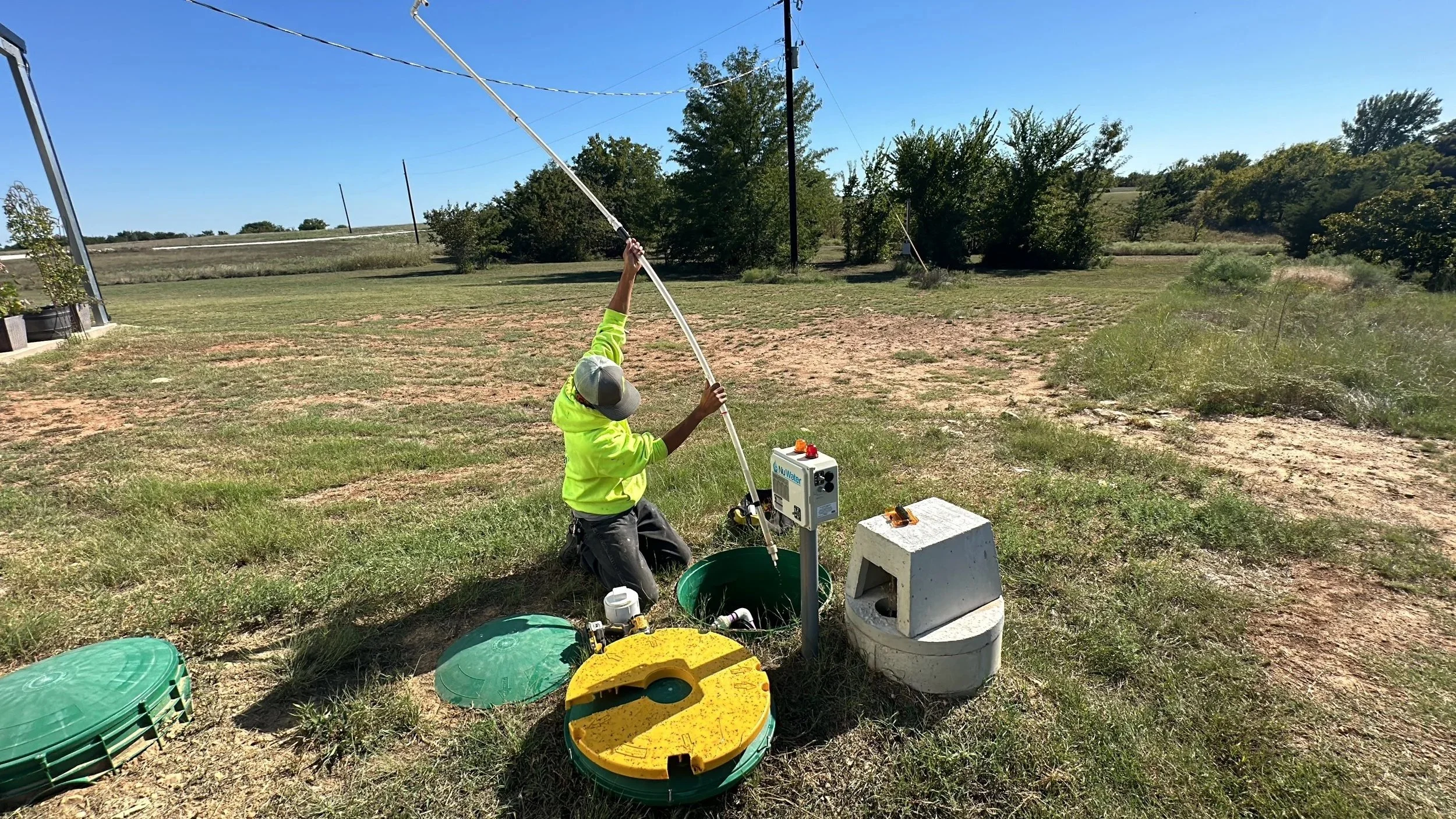 A person kneeling on the grass in a rural area, operating equipment from a water testing or well monitoring system, surrounded by green bins and utility boxes, under a clear blue sky.