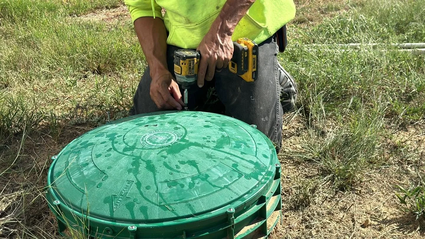 A person in a bright yellow-green shirt works outside with a drill near a green underground utility access cover in a grassy area.