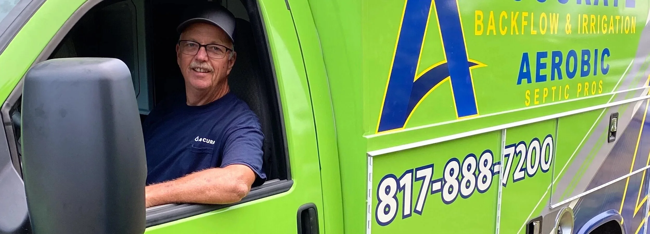A smiling man sitting in the driver's seat of a bright green service truck with company branding for backflow and irrigation services, including a phone number.